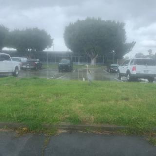A cloudy day at a parking lot with several parked cars and trees in the background.