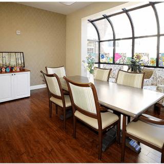 A well-lit dining room with a wooden table and cushioned chairs, featuring a large window and hardwood flooring.