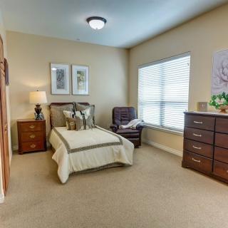 A cozy bedroom with a single bed, an armchair, and a chest of drawers, well-lit by a large window.