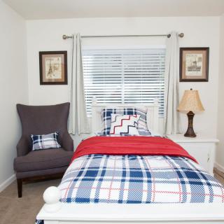A tidy bedroom with a single bed featuring plaid bedding, a chair, and a lamp by the window.