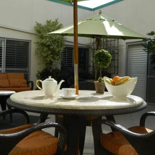 A cozy outdoor seating area featuring a round table with a tea set and a bowl of fresh fruit, shaded by an umbrella.