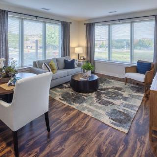 A modern living room with large windows, featuring a sofa, chairs, a wooden table, and a decorative rug.