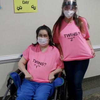 Two individuals wearing matching pink 'Twins?' shirts, one seated in a wheelchair and both wearing masks.