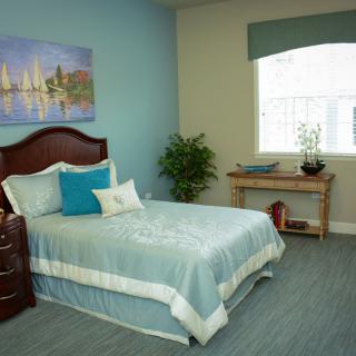 A cozy bedroom with a neatly made bed, decorative pillows, a potted plant, and a landscape painting on the wall.