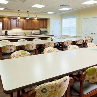 A modern community dining area with long tables, patterned chairs, and an open kitchen layout.