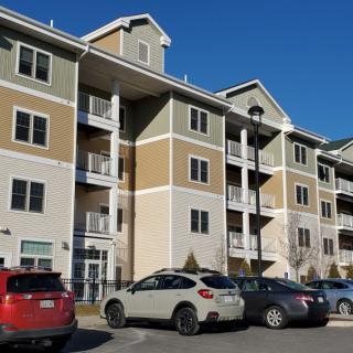 A modern apartment building with several parked cars in front under a clear blue sky.