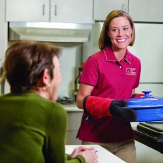 A woman in a red shirt with oven mitts serving a dish to another person in a kitchen setting.