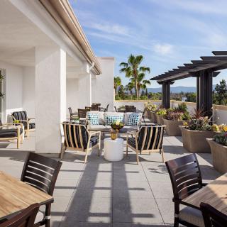 A sunny outdoor patio with tables and chairs, surrounded by plants.