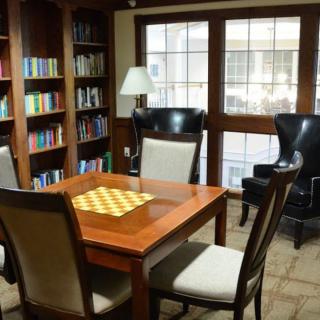 A cozy library reading room with bookshelves, a table with a chessboard, and comfortable seating near windows.