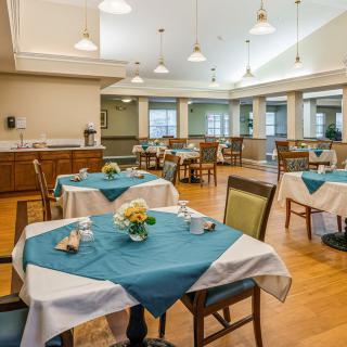 A well-lit dining room with neatly arranged tables and chairs, featuring blue and white tablecloths.