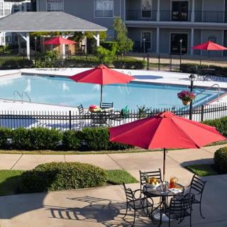 An outdoor swimming pool area with red umbrellas and patio furniture, surrounded by an apartment complex on a sunny day.