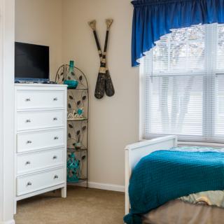 A cozy bedroom featuring a white dresser with a television on top, blue-accented decor, and a bed near a window with blue curtains.