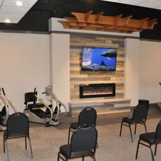 A fitness room with exercise equipment, chairs facing a television above a fireplace.