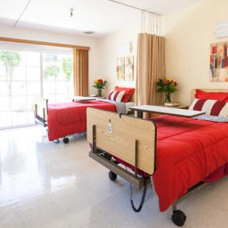 A well-lit hospital patient room featuring two beds with red bedding.