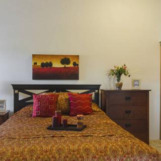 A cozy bedroom with warm-toned bedding and vibrant cushions, featuring a landscape painting above the bed and a dresser adorned with a vase of flowers.