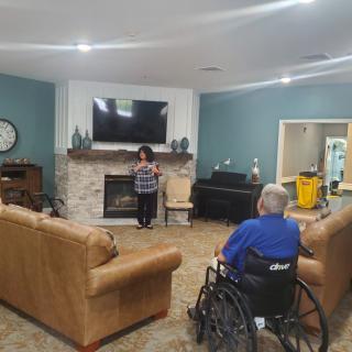 A person in a wheelchair faces someone standing in a living room with a fireplace and television.