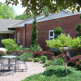 A serene garden patio with outdoor seating and lush greenery beside a brick building.