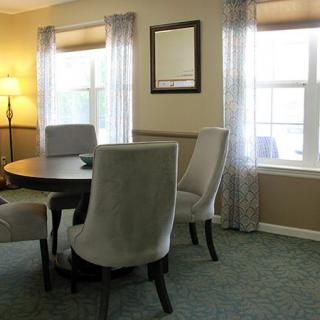A cozy dining room with a round table and upholstered chairs, featuring large windows and soft lighting.