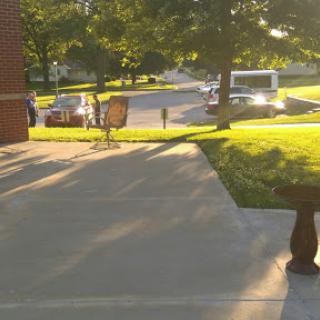 A sunlit view of a sidewalk leading to a street with parked vehicles and trees in the background.