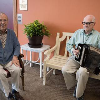 Two elderly men, one playing an accordion and the other seated beside him, enjoy a moment indoors next to a potted plant.