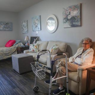 An elderly woman sitting in a cozy living room with a sofa and walker nearby.