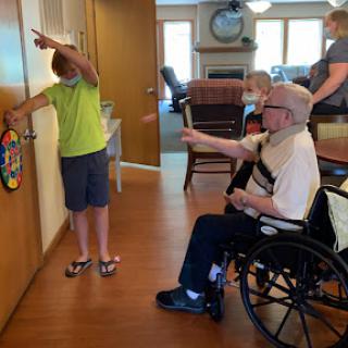 A child engages with an elderly person in a care facility, pointing at a door while another staff member observes.
