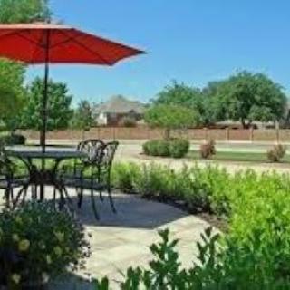 A sunny patio with a table and chairs under a red umbrella, surrounded by greenery.