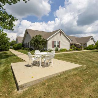 A suburban house with a small patio area featuring outdoor furniture on a grassy lawn under a cloudy sky.