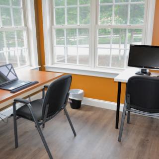 A bright office workspace featuring desks, computers, and chairs near large windows.