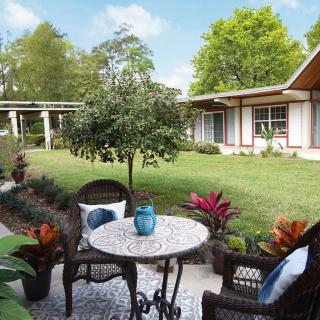 A serene garden patio featuring wicker chairs and a round table set against a backdrop of lush greenery.