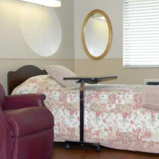 A bedroom with a hospital bed featuring floral bedding, a maroon armchair, and a wall-mounted mirror next to a window.