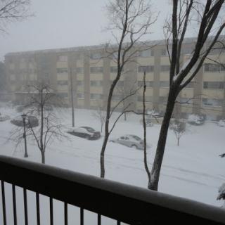 A view of a snow-covered street and apartment building during a winter storm.