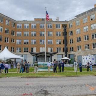 A community gathering in front of a historic building under a cloudy sky, featuring a flagpole and attendees enjoying the event.