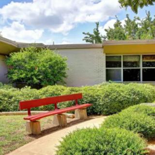 A tranquil outdoor scene featuring a red bench surrounded by greenery and a modern building in the background.
