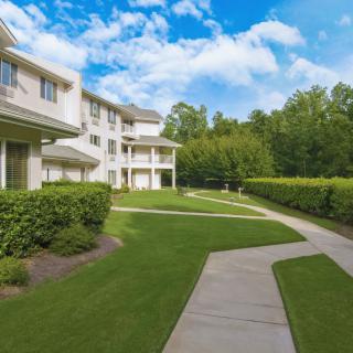 A residential building with a well-maintained lawn and sidewalk, surrounded by trees under a partly cloudy sky.