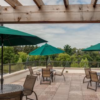 A rooftop patio with tables, chairs, and green umbrellas under a pergola overlooking lush greenery.
