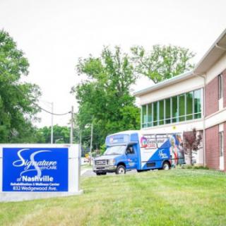 A building with a sign and a parked van on a grassy area, surrounded by trees.