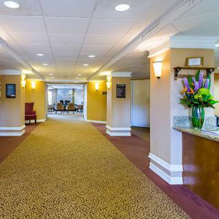 A brightly lit hotel lobby with a carpeted hallway, reception area, and decorative flowers.