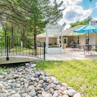 A backyard with a patio featuring outdoor furniture and an umbrella, a small bridge over rocks, surrounded by trees.