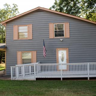 A two-story house with wooden siding, featuring a front porch and an American flag, surrounded by trees.