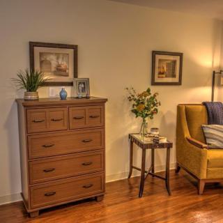 A cozy living room corner featuring a wooden dresser, framed pictures, and a yellow armchair.