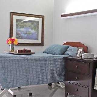 A hospital room with a neatly made bed, a nightstand holding flowers and books, and a framed painting on the wall.