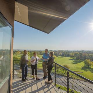 A group of people stand on a balcony overlooking a golf course on a sunny day.