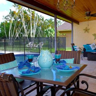 A cozy outdoor patio with a dining table set and blue decor next to a pool, surrounded by lush greenery.