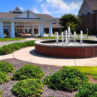 A scenic view of a fountain surrounded by greenery in front of a building on a sunny day.