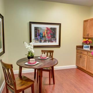 A cozy kitchenette with a round table set for two, featuring wooden chairs, a decorative orchid centerpiece, and framed artwork on a light green wall.