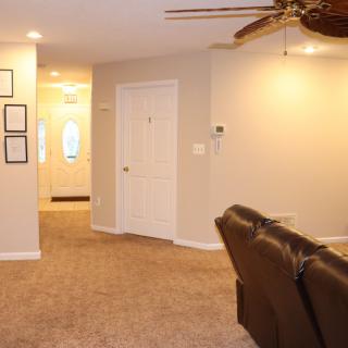A cozy living room with carpet flooring, a ceiling fan, and a comfortable recliner leading to a bright hallway.
