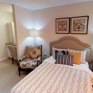A cozy bedroom with a neatly made bed, decorative pillows, and a patterned armchair beside a floor lamp.