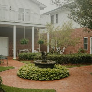 A serene courtyard featuring a central fountain, surrounded by lush greenery and a brick pathway.