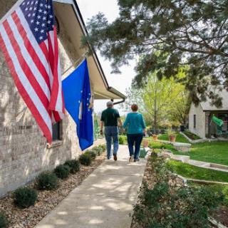 A couple walking along a pathway next to their home, adorned with American and state flags, surrounded by lush greenery.
